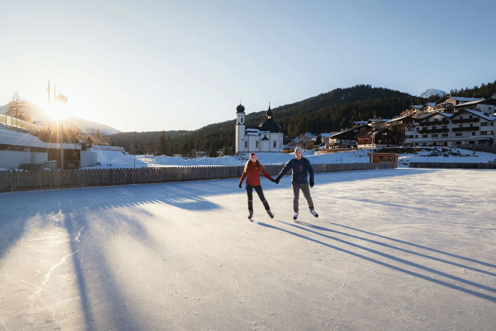 Two skaters crossing the ice of the Seefeld rink beside the Congress Centre in winter sun, with a white chapel with an onion dome and hotels along the edge. — Eislaufplatz Kongresszentrum Seefeld