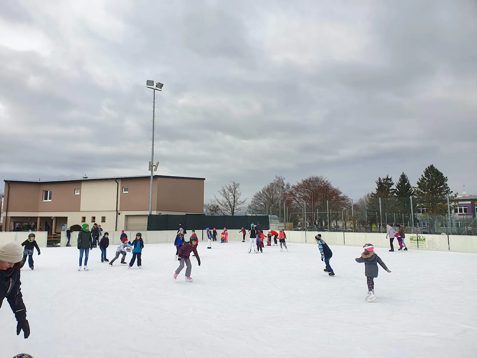 Skaters on the open-air rink at Leonhofen under floodlights and a grey winter sky, with the club house behind the boards – Eislaufplatz Leonhofen