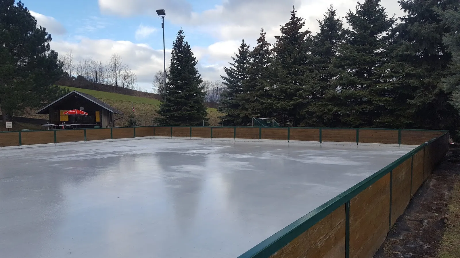 Das glatte, leere Eis der Eislaufbahn Markt Piesting mit Holzbande, einem kleinen Standl und einer Reihe von Fichten dahinter. – Eislaufplatz Markt Piesting