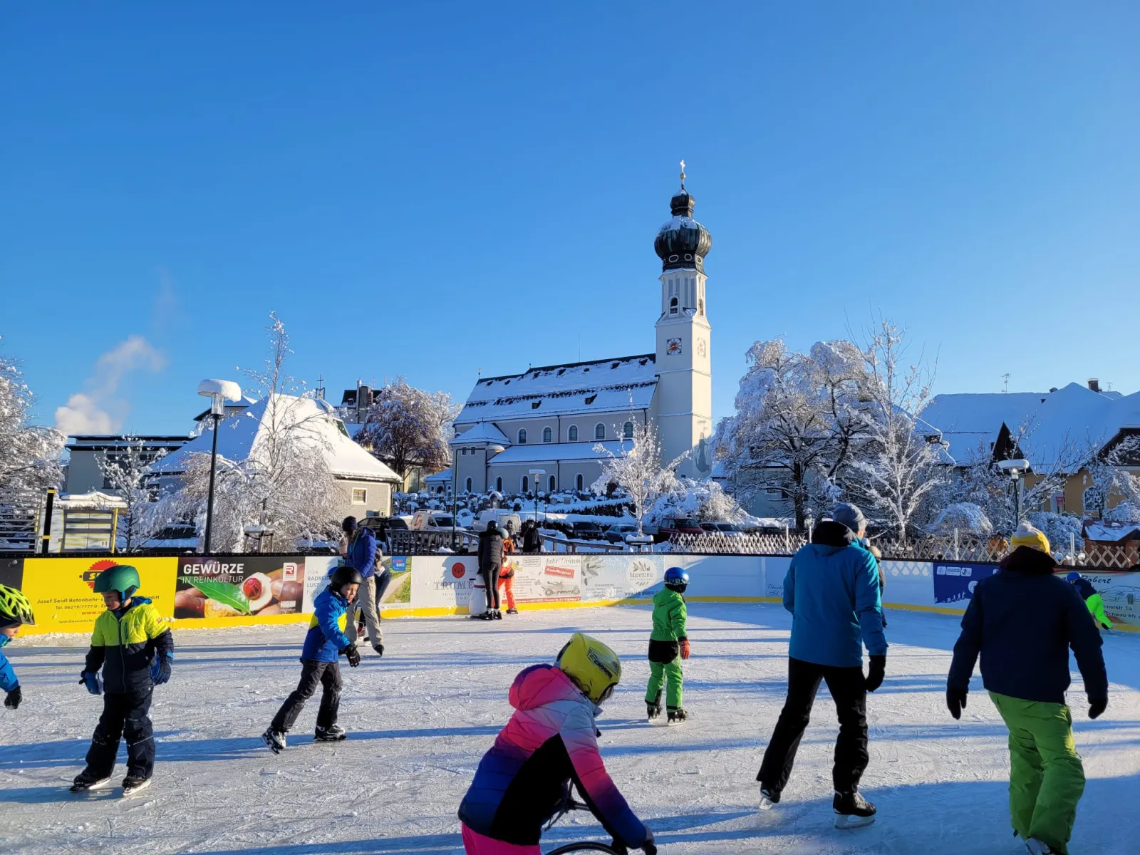 Children and adults skating on the snow-edged rink in Obertrum am See, with the church and the village houses behind. — Eislaufplatz Obertrum am See