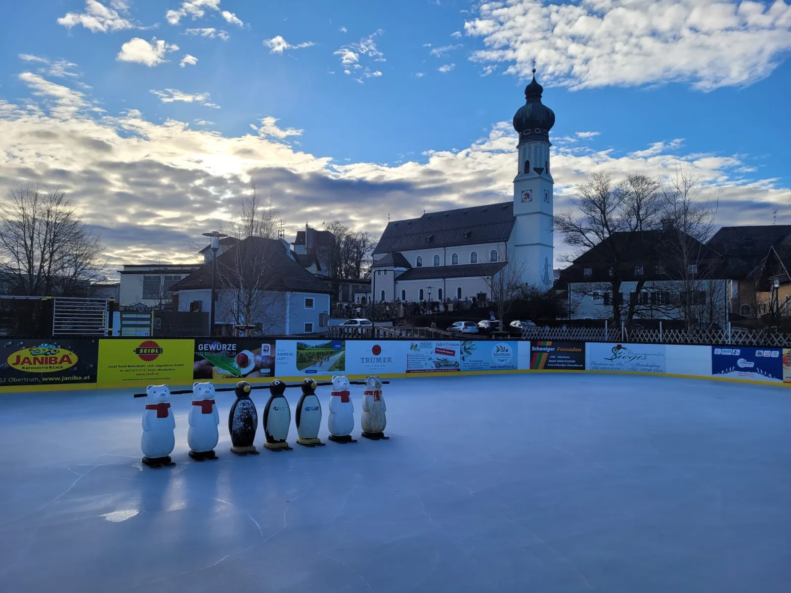 The empty rink in Obertrum am See at sunset, with a row of penguin skating aids along the sponsor boards and the parish church tower behind. — Eislaufplatz Obertrum am See
