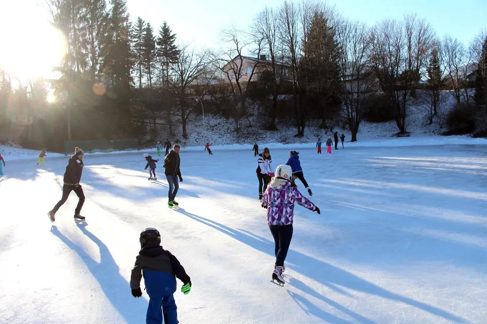 Skaters on the sunny Pöls-Oberkurzheim ice rink in winter, with long shadows and bare trees backlit by the low sun. — Eislaufplatz Pöls-Oberkurzheim