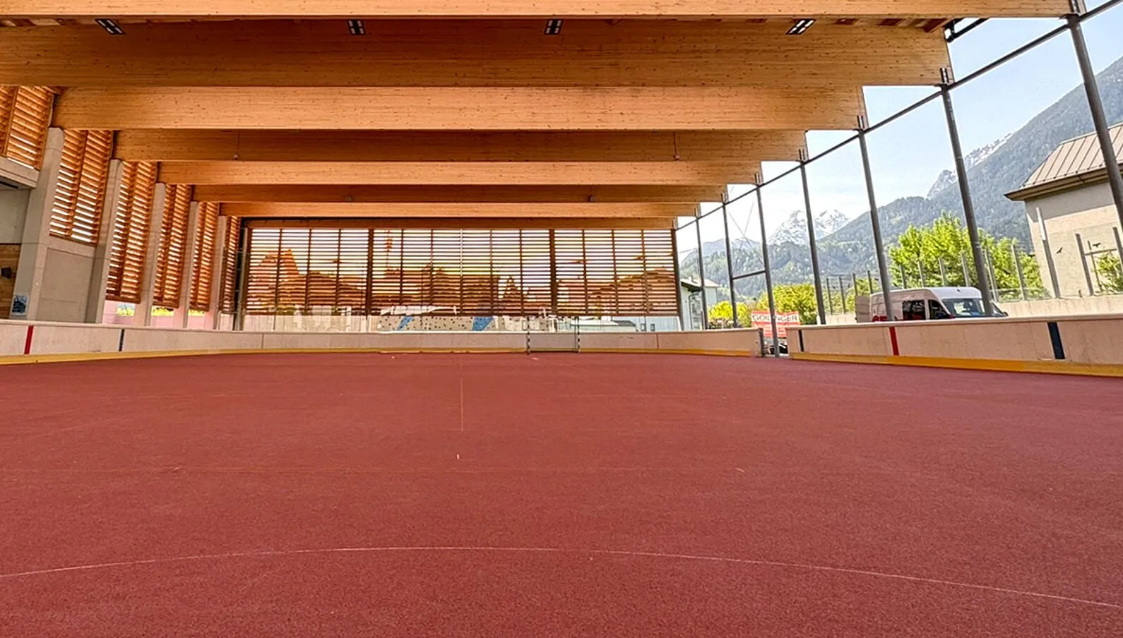The timber-roofed, open-sided hall over the Schwaz rink in summer, without ice: red rubber floor, boards, wooden roof beams and the mountains beyond the glass side. — Eislaufplatz Schwaz