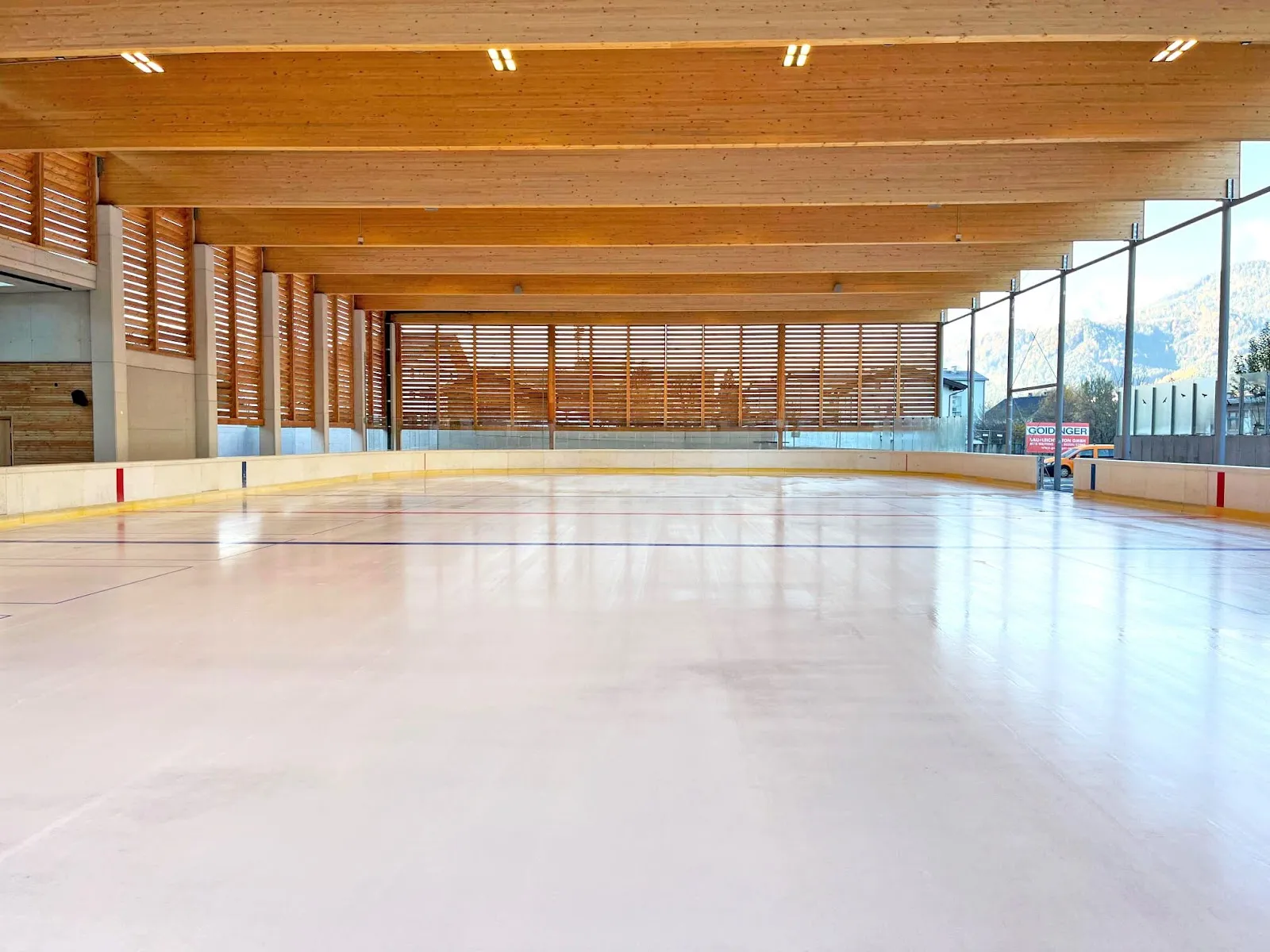 The empty ice surface under the open-sided timber roof of the Kaltlufthalle, with the boards and the mountains visible through the side. — Eislaufplatz Schwaz