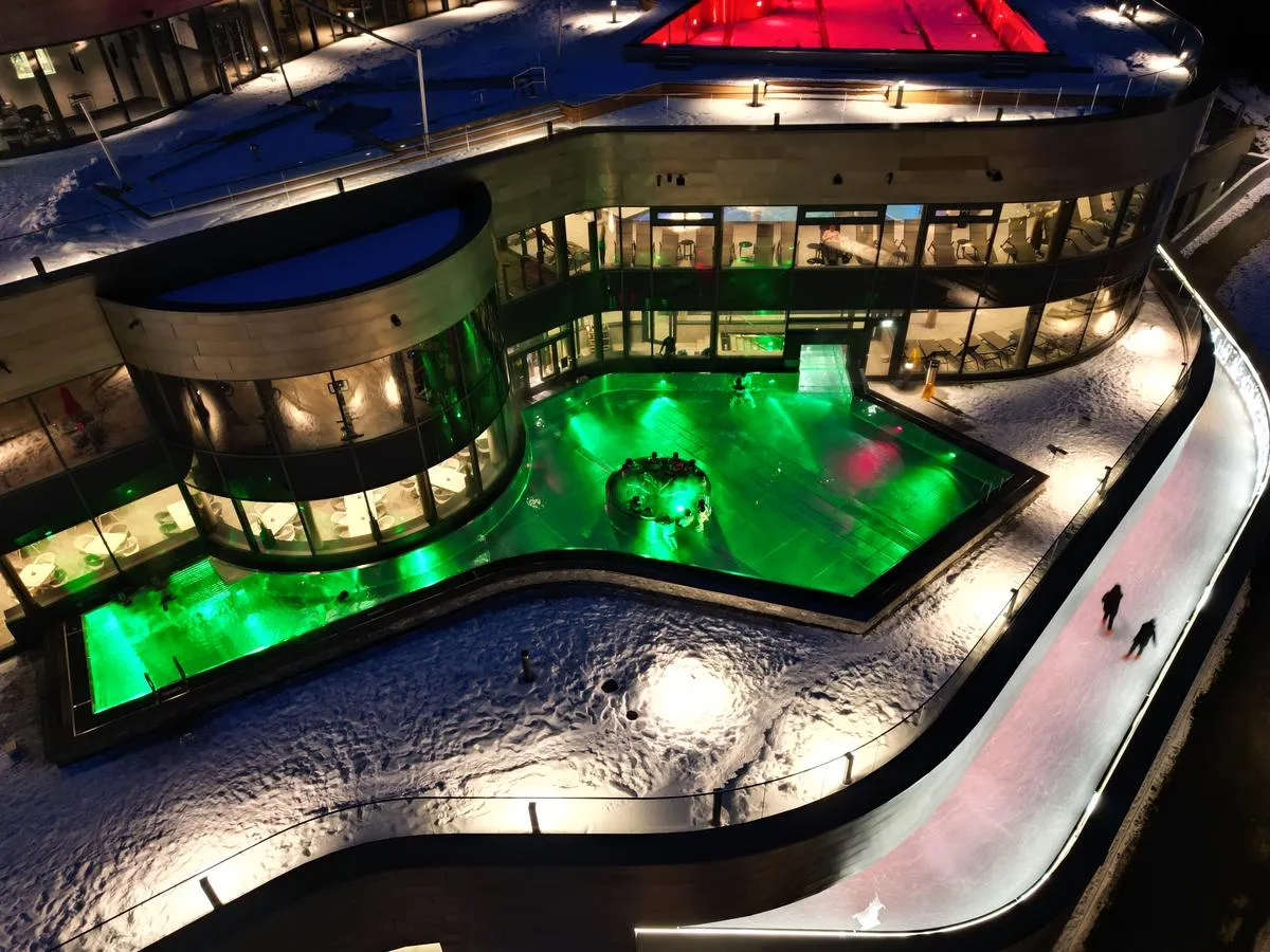 Night view from above of the Silvretta Therme in Ischgl, with the lit ice track curving around the building on the right and two skaters on it. – Eislaufplatz Silvretta Therme Ischgl