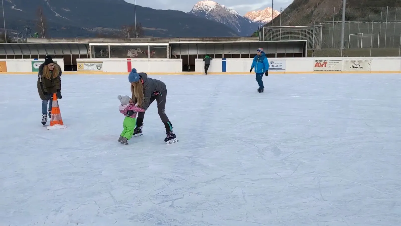 The pale ice surface of the Imst open-air rink with a child skating beside the boards and the sports centre roof behind. — Eislaufplatz Sportzentrum Imst