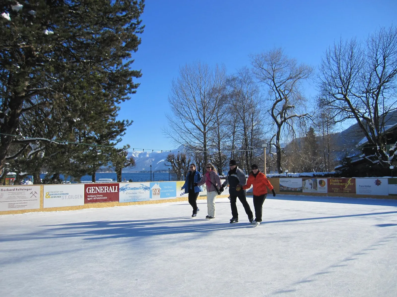 A group of skaters holding hands on the St. Gilgen rink on a sunny day, with sponsor boards, bare trees and distant hills behind. — Eislaufplatz St. Gilgen