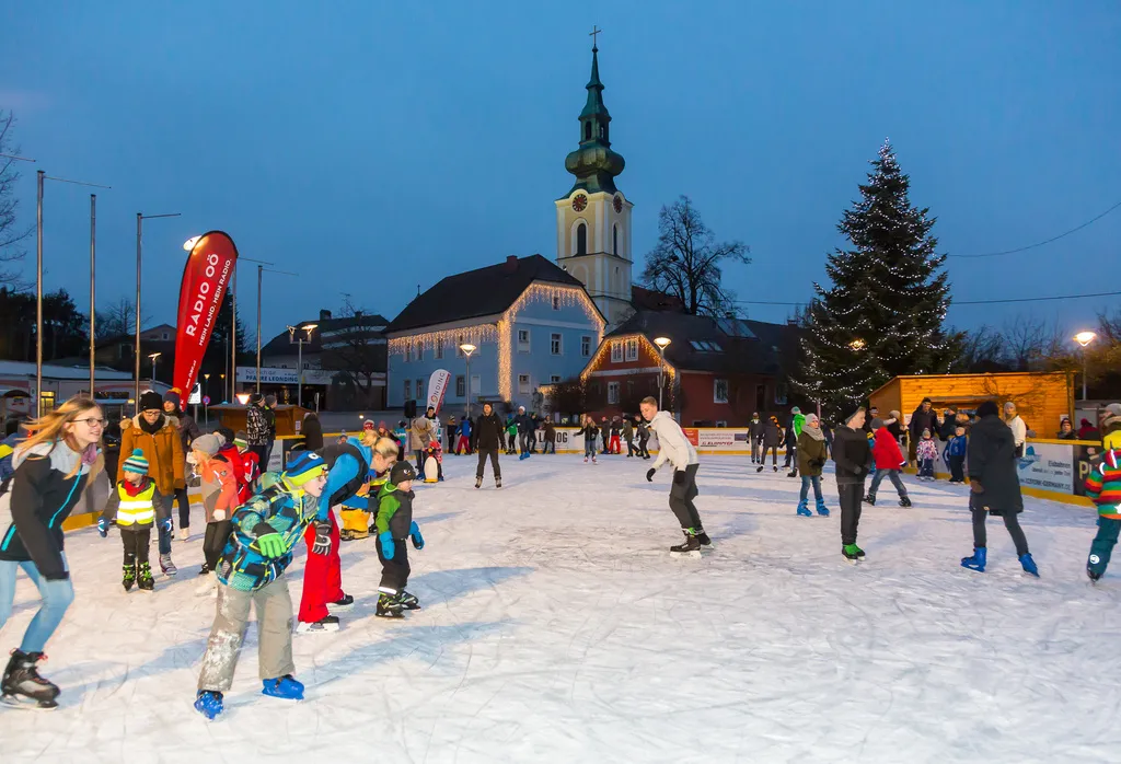 The rink at dusk on the Stadtplatz, with skaters across the ice, a Christmas tree and the church tower in the background. — Eislaufplatz Stadtplatz
