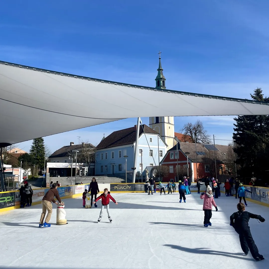Skaters on the open-air ice on the Stadtplatz under a white sail canopy, with the church tower and the town's old houses behind. — Eislaufplatz Stadtplatz