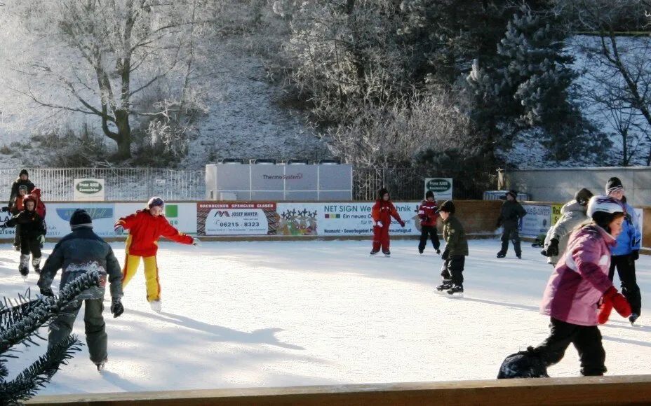 Children skating on the refrigerated Straßwalchen rink in winter sun, with the chiller unit and sponsor boards along the far boards and frosted trees behind. — Eislaufplatz Straßwalchen
