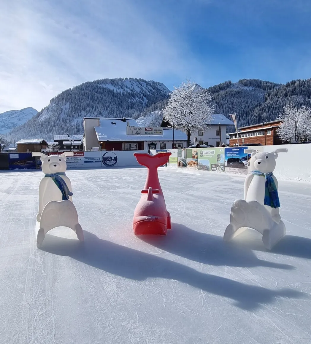 The ice surface of the Tannheim ice rink on a sunny winter day, with two white polar-bear skating aids and a red whale in the middle, advertising boards behind and snow-covered mountains above the village. — Eislaufplatz Tannheim