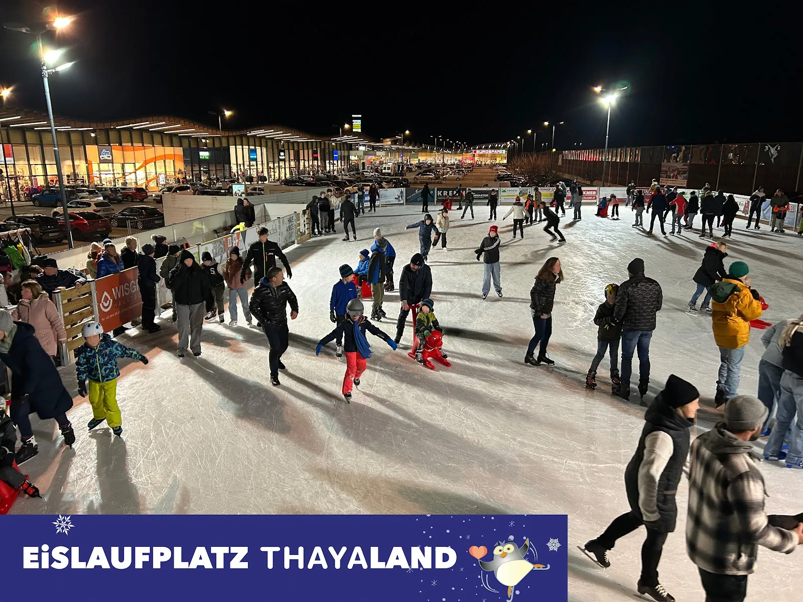 A busy evening at the floodlit Thayaland open-air rink with skaters across the ice and the park buildings lit behind. – Eislaufplatz Thayaland