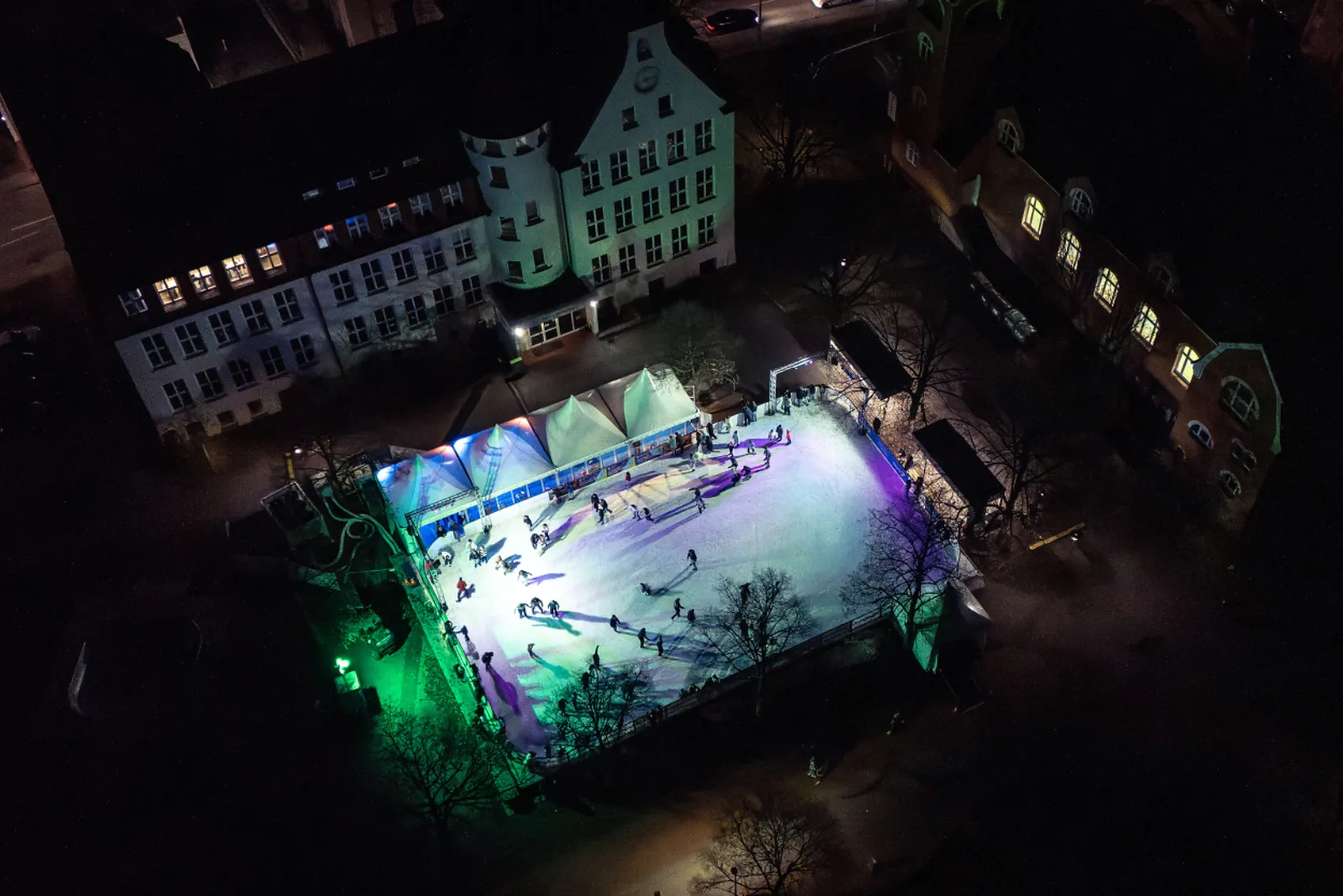 Nächtliche Luftaufnahme der mobilen Eisbahn auf dem Bohlschulplatz in Aalen, mit Schlittschuhläufern auf dem beleuchteten Eis unter weißen Zeltdächern und dem alten Schulgebäude dahinter. – Eispark Aalen