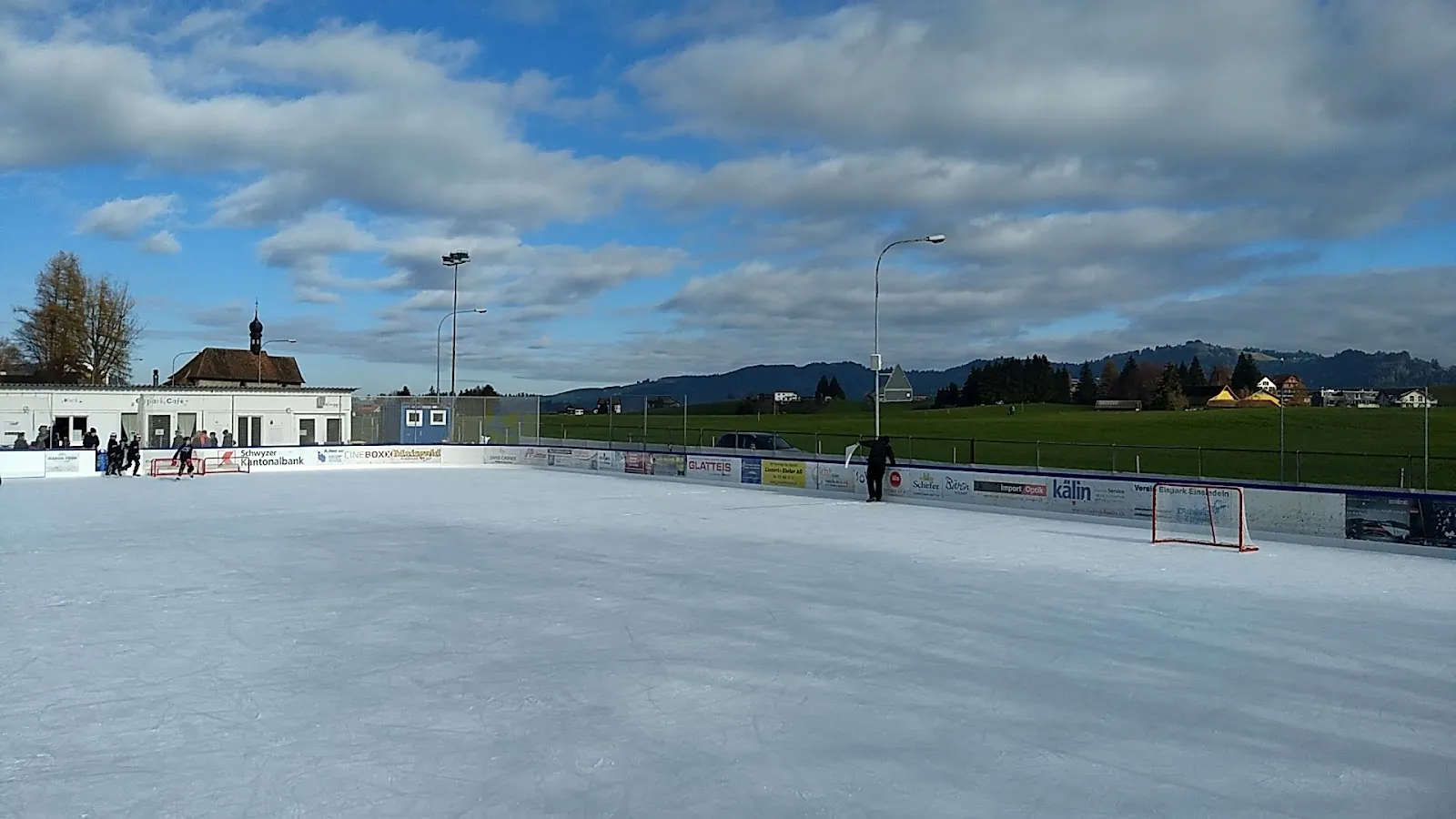 The open-air Eispark Einsiedeln ice under a blue sky with the clubhouse and the rink boards in the distance – Eispark Einsiedeln
