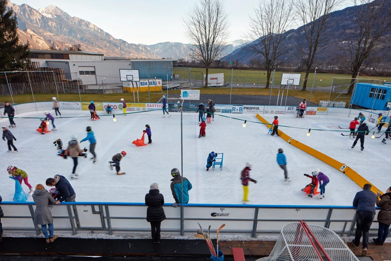 Läuferinnen und Läufer verteilen sich auf dem Freiluft-Eis, am Rand eine gelbe Bande und hinter der Sportanlage die Alpengipfel. – Eispark Sarganserland