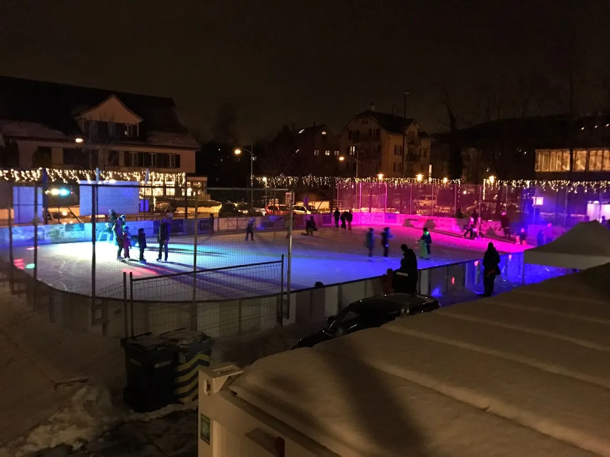 Das beleuchtete Eisfeld des Eisparks Uetikon, nachts von ausserhalb seines Zauns gesehen, mit den Dorfhäusern dahinter – Eispark Uetikon
