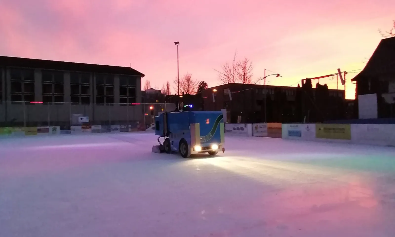 Das Eisfeld des Eisparks Uetikon in der Dämmerung unter rosa Himmel, mit einer Eisaufbereitungsmaschine auf dem Eis und den Schulgebäuden dahinter – Eispark Uetikon