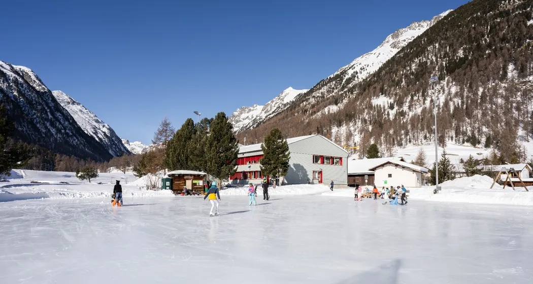 Skaters spread across the natural-ice field at Bever, with the village and snowy peaks of the Engadin behind. — Eisplatz Bever
