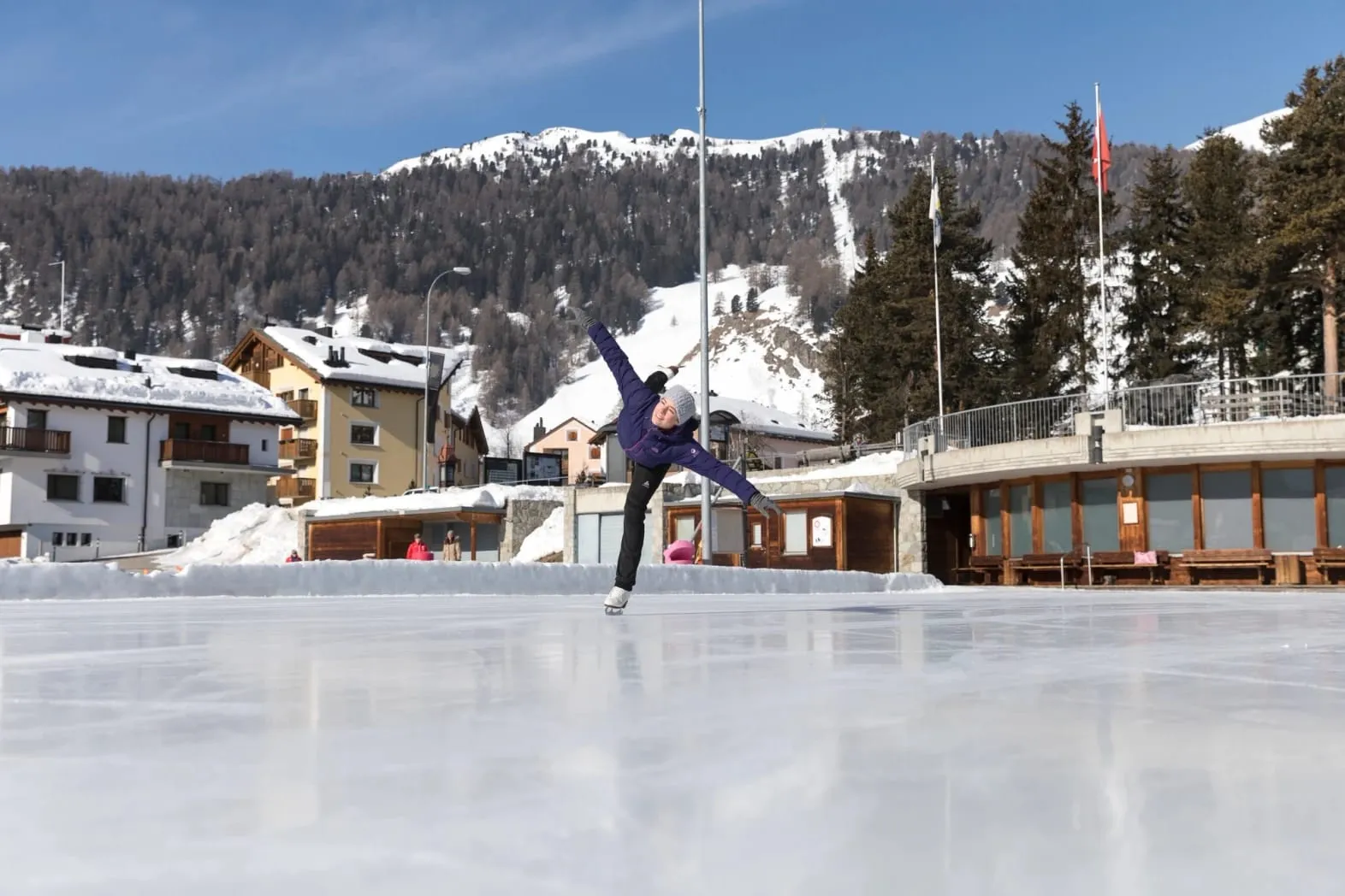 A skater on the smooth ice of the Celerina Center da Sport rink, with the sports centre buildings and snowy hills behind. — Eisplatz Celerina Center da Sport