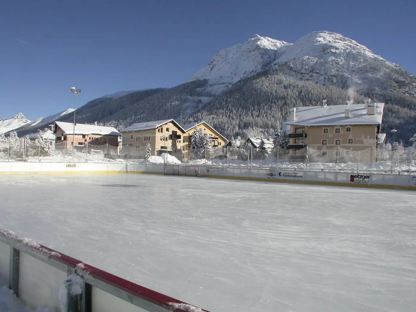 The ice of the La Punt Chamues-ch rink with the board edge in the foreground, the village rooftops and a snowy peak behind. — Eisplatz La Punt