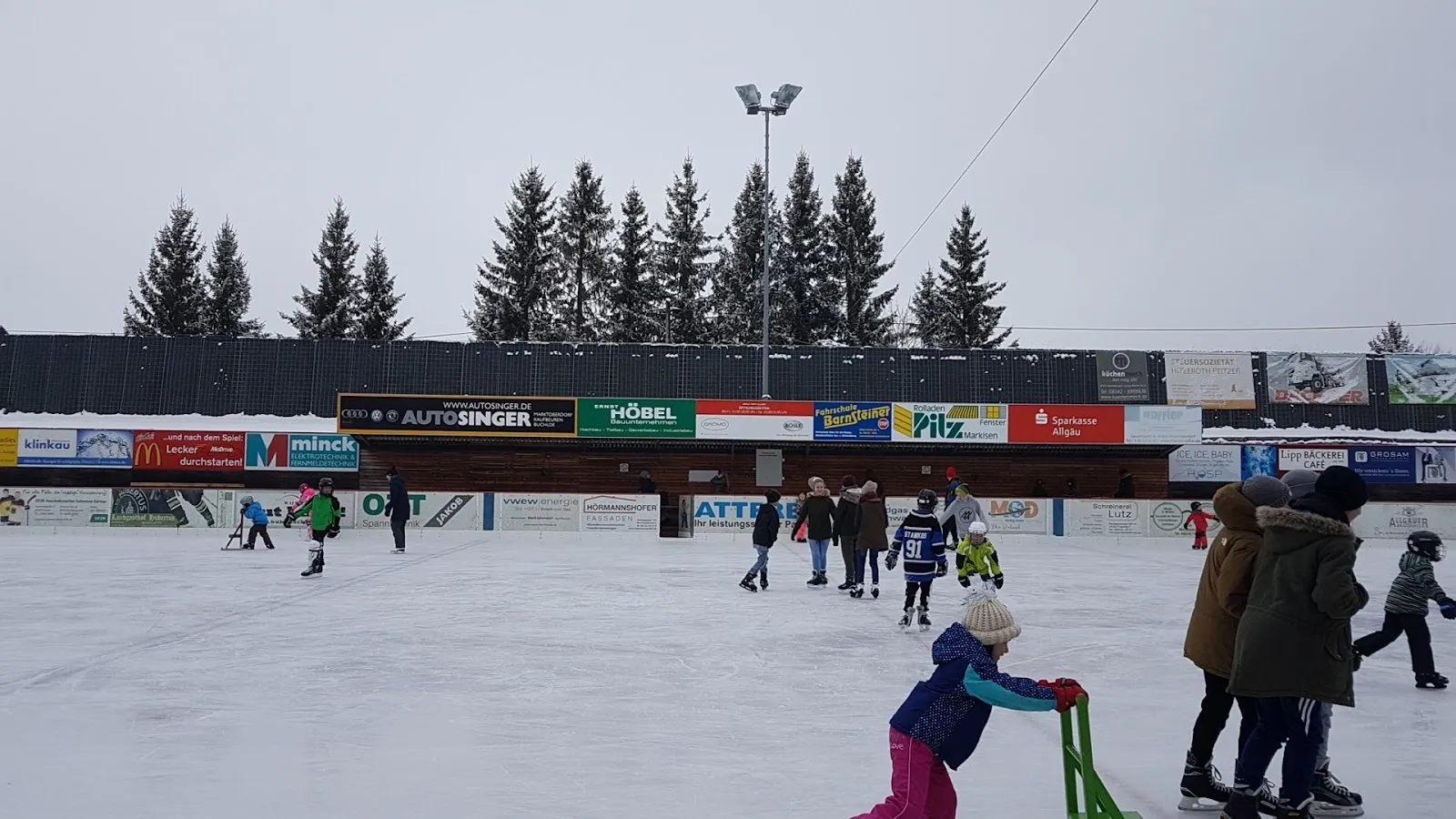 The open-air Eisplatz in Marktoberdorf under grey winter skies, with skaters on the ice and the pine trees and rink-side building behind. — Eisplatz Marktoberdorf