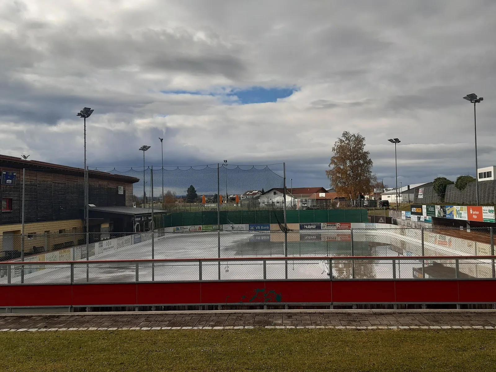 The open-air rink seen across its red boards on a bright afternoon, a nearly empty ice sheet with floodlight masts and the timber-clad clubhouse at the far side. — Eisplatz Marktoberdorf