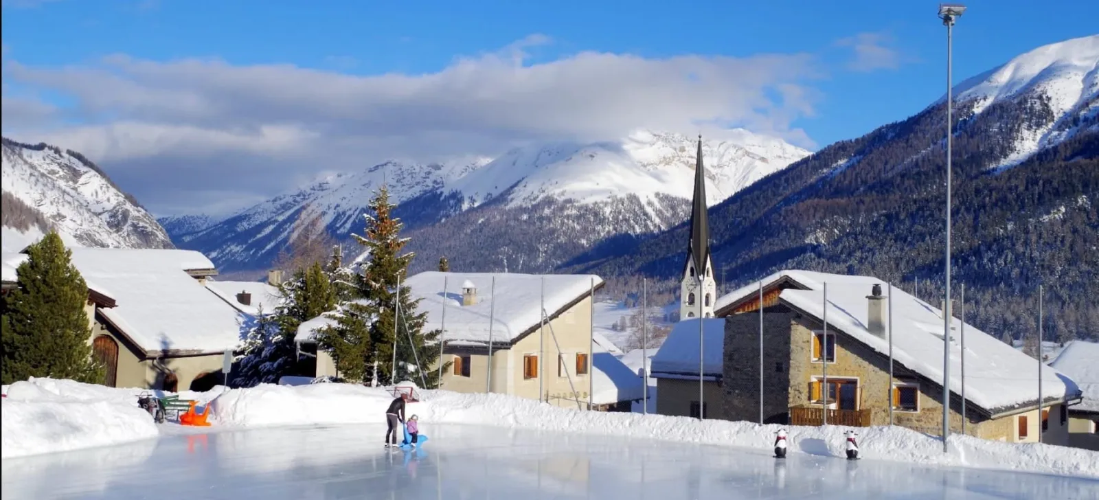 The Purtum ice field at Zuoz with a skater in the distance, the village church tower and snowy mountains behind. — Eisplatz Purtum Zuoz