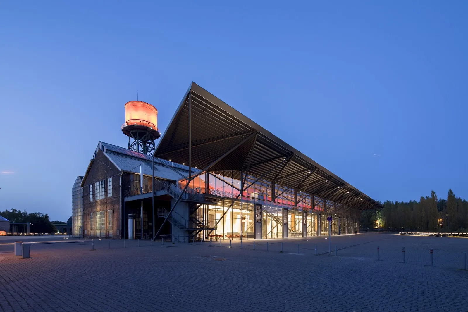 Die Jahrhunderthalle in Bochum in der Dämmerung, die Industriehalle mit ihrem schrägen Dach und dem beleuchteten Eingang, in der der EisSalon Ruhr aufgebaut ist. – EisSalon Ruhr