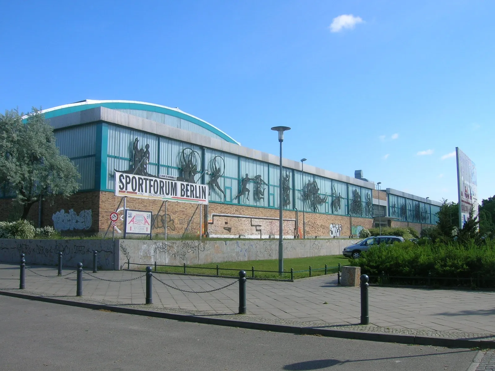 The long glazed and brick front of the Sportforum Berlin speed-skating hall with its curved roof, seen from the pavement opposite. — Eisschnelllaufhalle Berlin-Hohenschönhausen