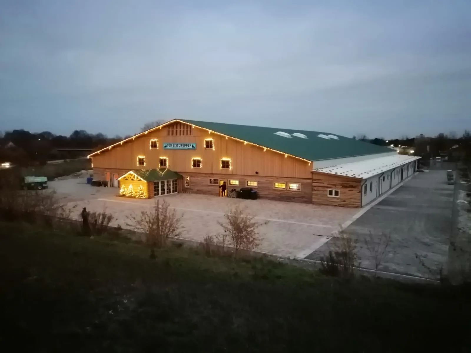 Die holzverkleidete Halle des Eisschuppens Lippstadt mit ihren beleuchteten Fenstern in der Abenddämmerung, über den Kiesparkplatz hinweg gesehen – Der Eisschuppen Lippstadt