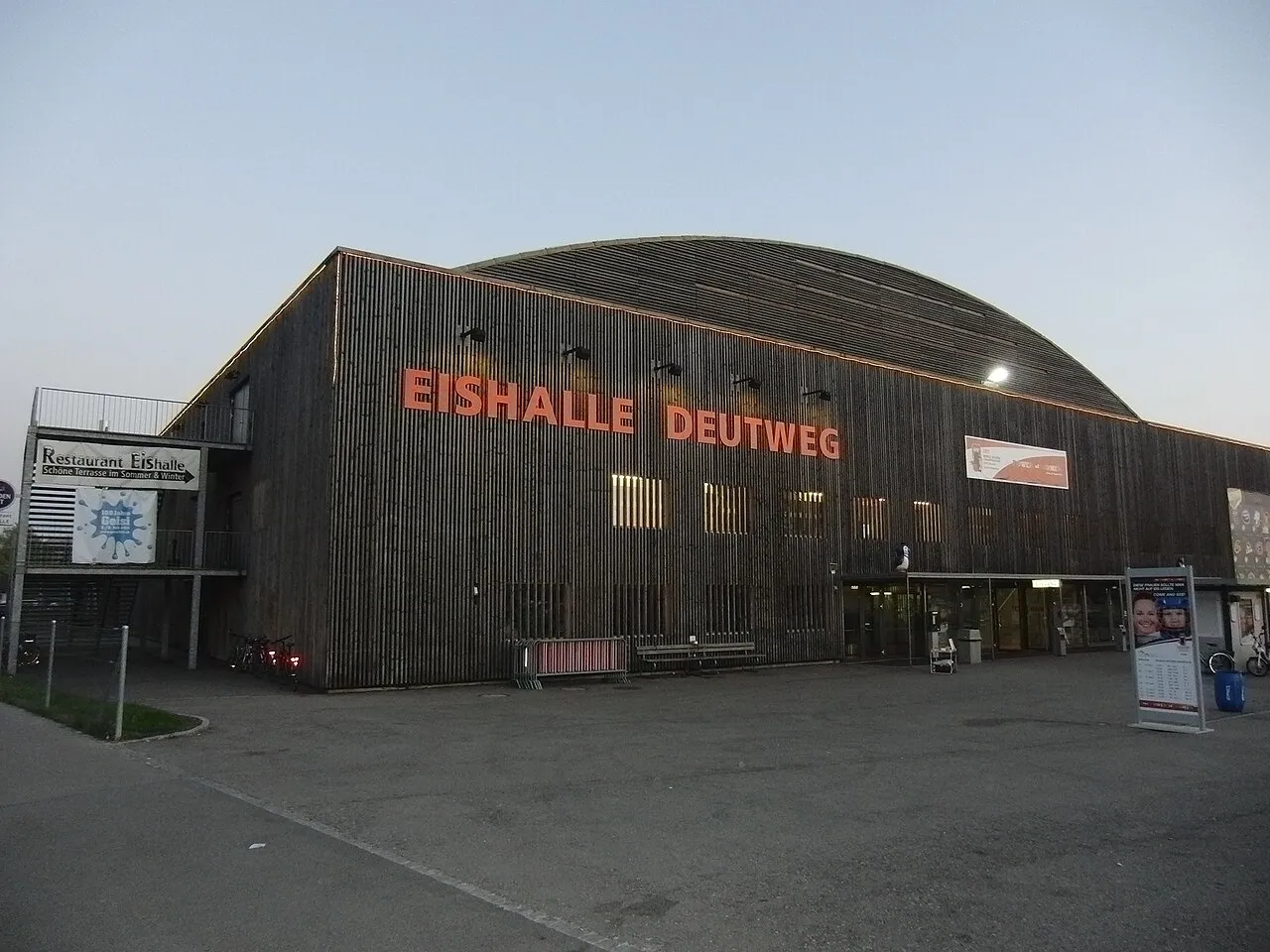 The wooden barrel-roofed Eishalle Deutweg in Winterthur seen from the forecourt at dusk, with its lit name lettering — Eissportanlage Deutweg