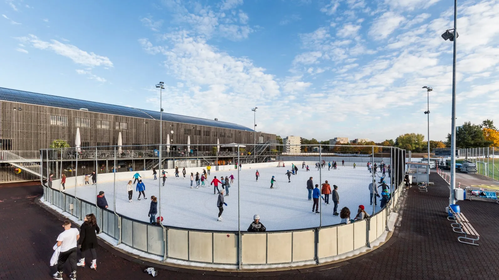 Skaters on one of the two outdoor ice fields of the Eissportanlage Deutweg in Winterthur, with the hall's timber-clad facade behind — Eissportanlage Deutweg