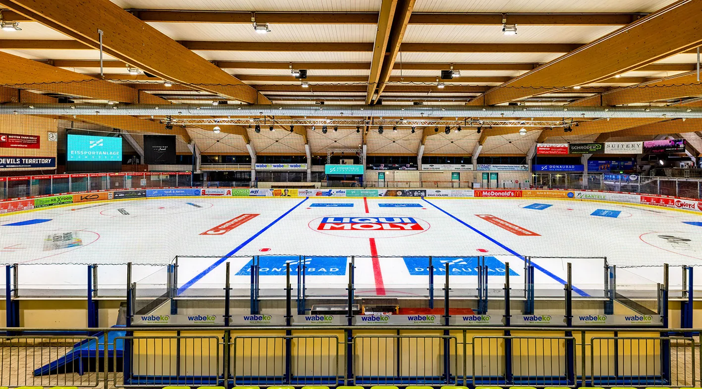 The empty indoor ice sheet with its blue and red hockey lines and Donaubad logos, surrounded by boards with sponsor panels under a laminated timber roof. — Eissportanlage Donaubad