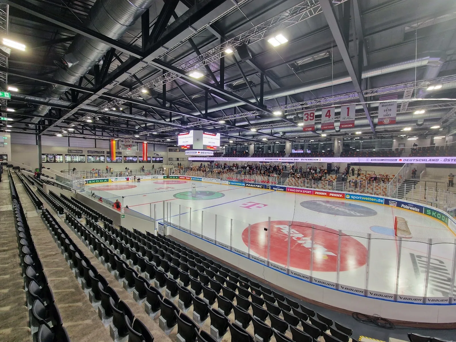 The ice sheet of the Landshut ice arena (VR-Bank Landshut Arena) seen from the stands, with the red centre-ice logo, the black seating tiers, the roof lighting and the scoreboard. — Städtische Eissportanlage Landshut
