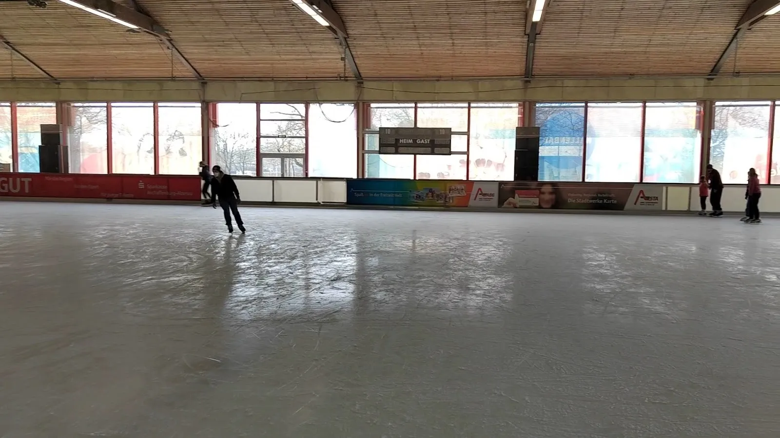 Die Eisfläche, über die Bande hinweg gesehen, mit einem einzelnen Läufer auf der gegenüberliegenden Seite, Werbebanden und einem Fensterband unter dem Holzdach. – Eissporthalle Aschaffenburg