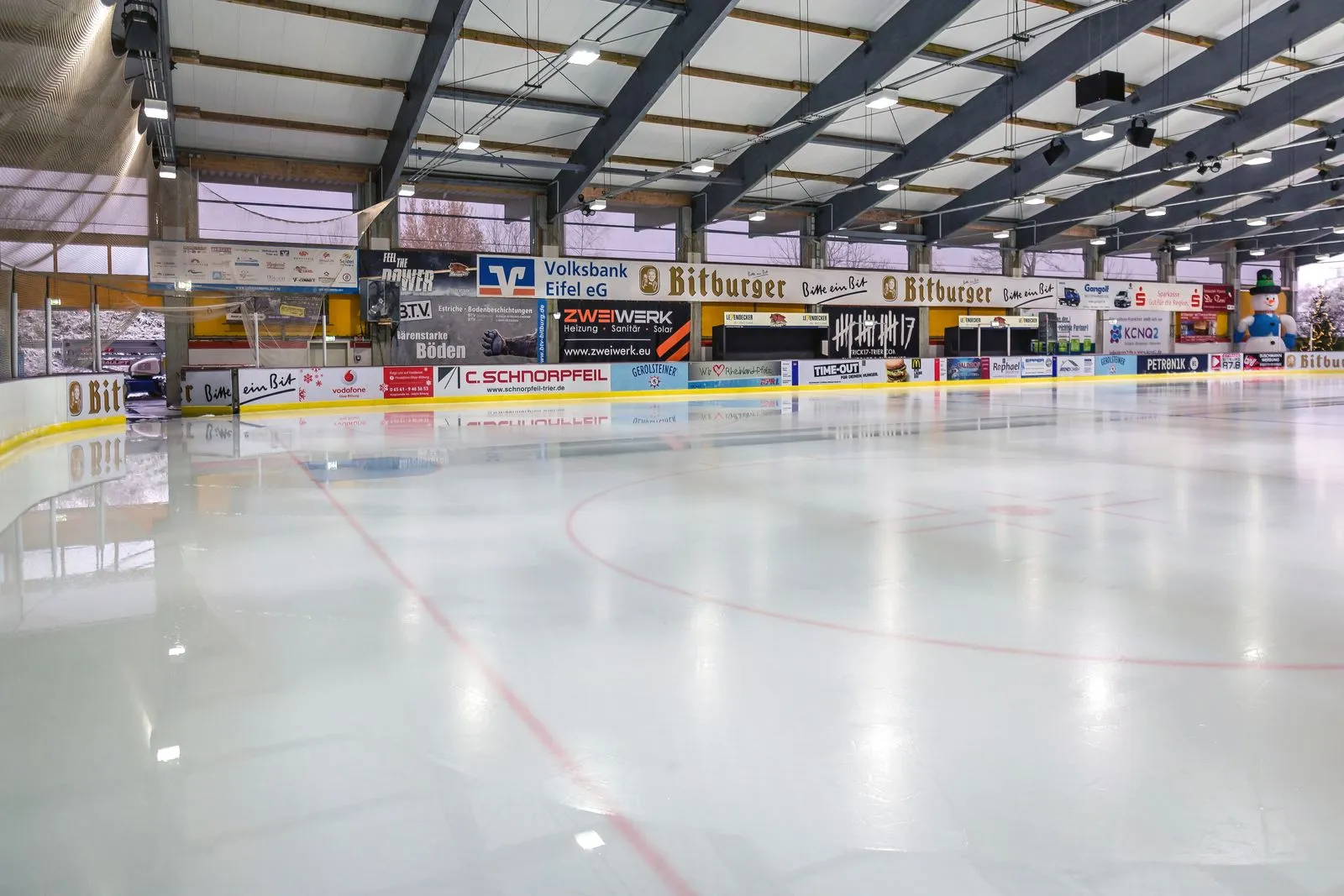 Die Eisfläche mit Banden, Werbetafeln und Stahlfachwerkdach, ohne Schlittschuhläufer auf dem Eis. – Eissporthalle Bitburg