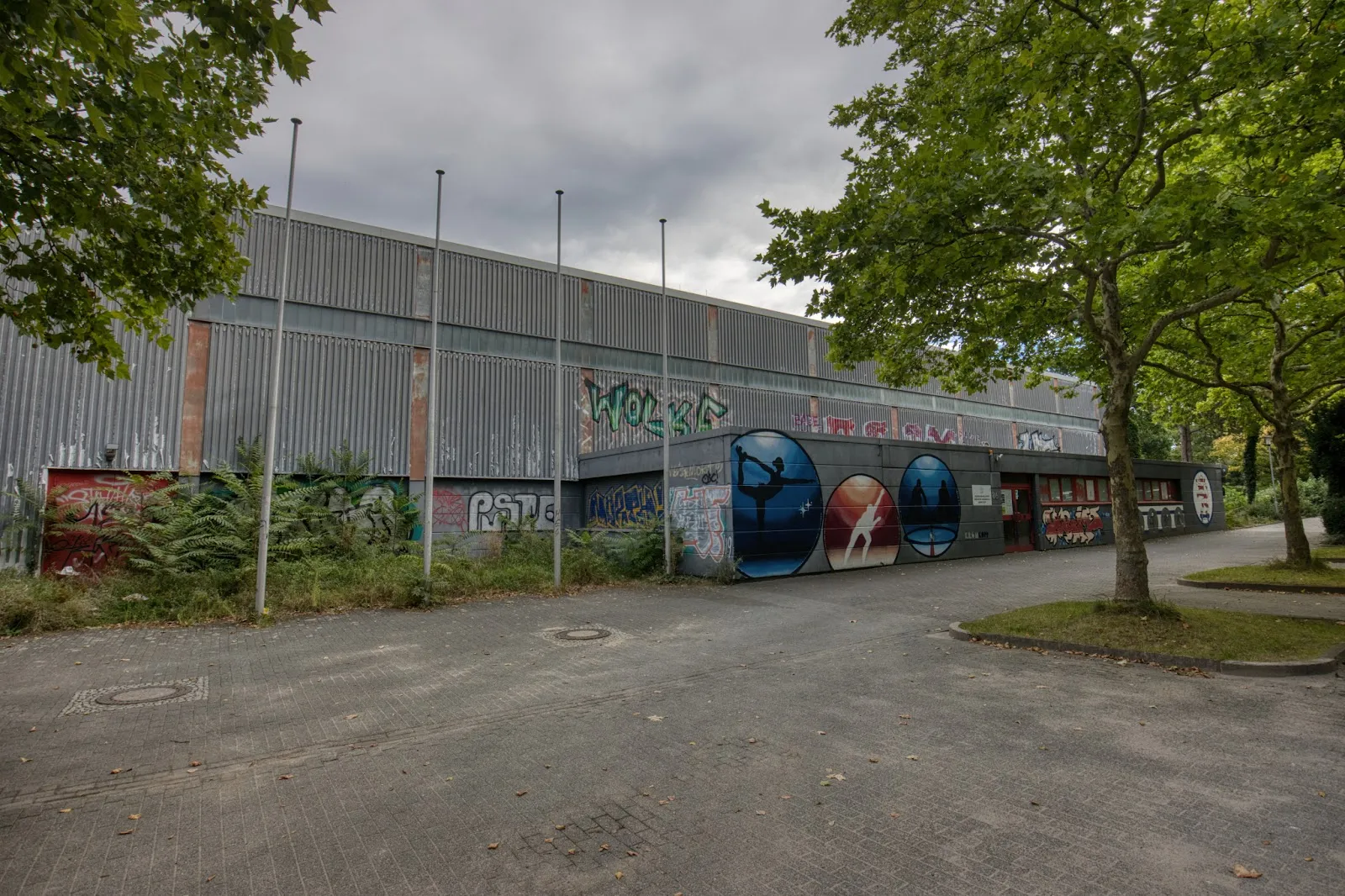 The side of the Darmstadt ice hall seen from a path under trees, with its grey metal cladding, flagpoles and painted panels. — Eissporthalle Darmstadt