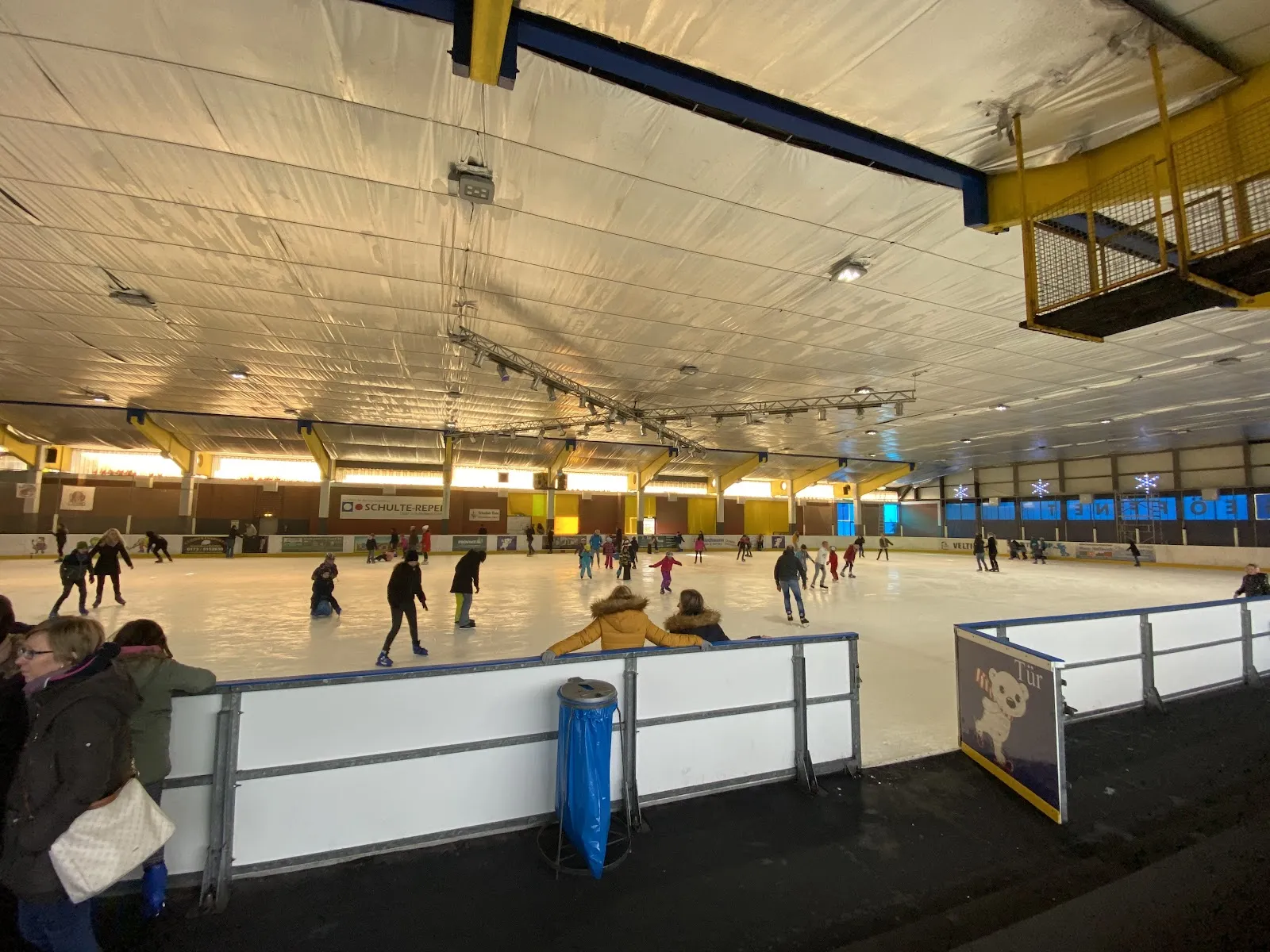 Schlittschuhläufer verteilt auf dem weißen Eis der Dorstener Eishalle unter dem hellen, zeltartigen Dach, mit gelben Stahlträgern und der Bande im Vordergrund. – Eissporthalle Dorsten