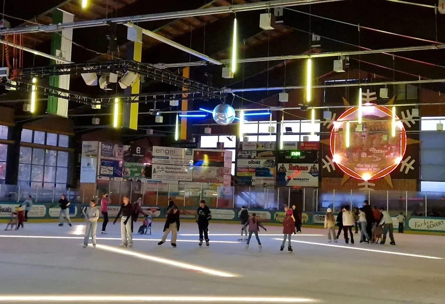 Schlittschuhläufer queren das Eis der Halle Herrischried unter bunten Partylichtern, darüber das Holz-Stahl-Dach. – Eissporthalle Herrischried