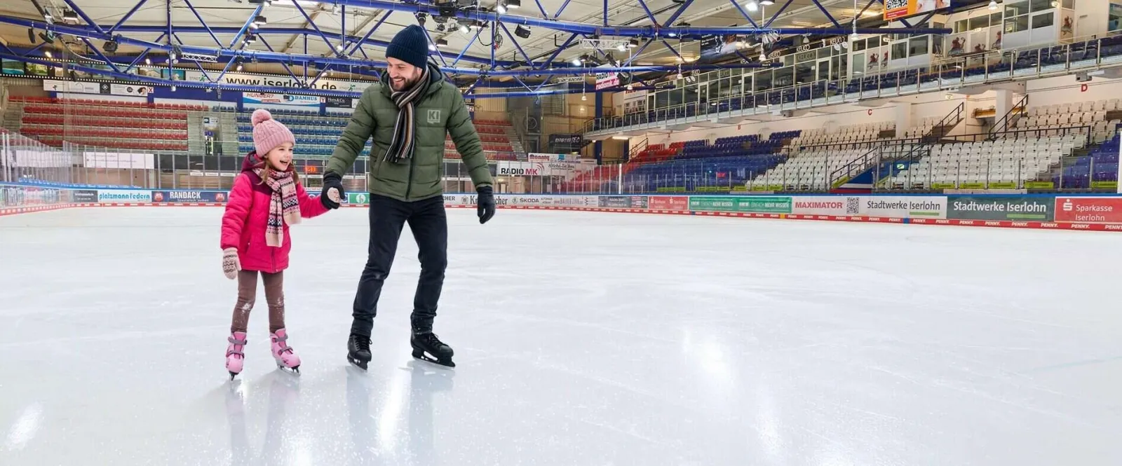 Zwei Schlittschuhläufer queren das weite weiße Eis der Halle Iserlohn, dahinter Tribünen und Dach. – Eissporthalle Iserlohn