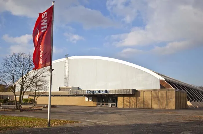 The Eissporthalle Neuss-Südpark seen from the car park: a white barrel-roofed hall with a brown brick entrance block and a tall banner flag. — Eissporthalle Neuss