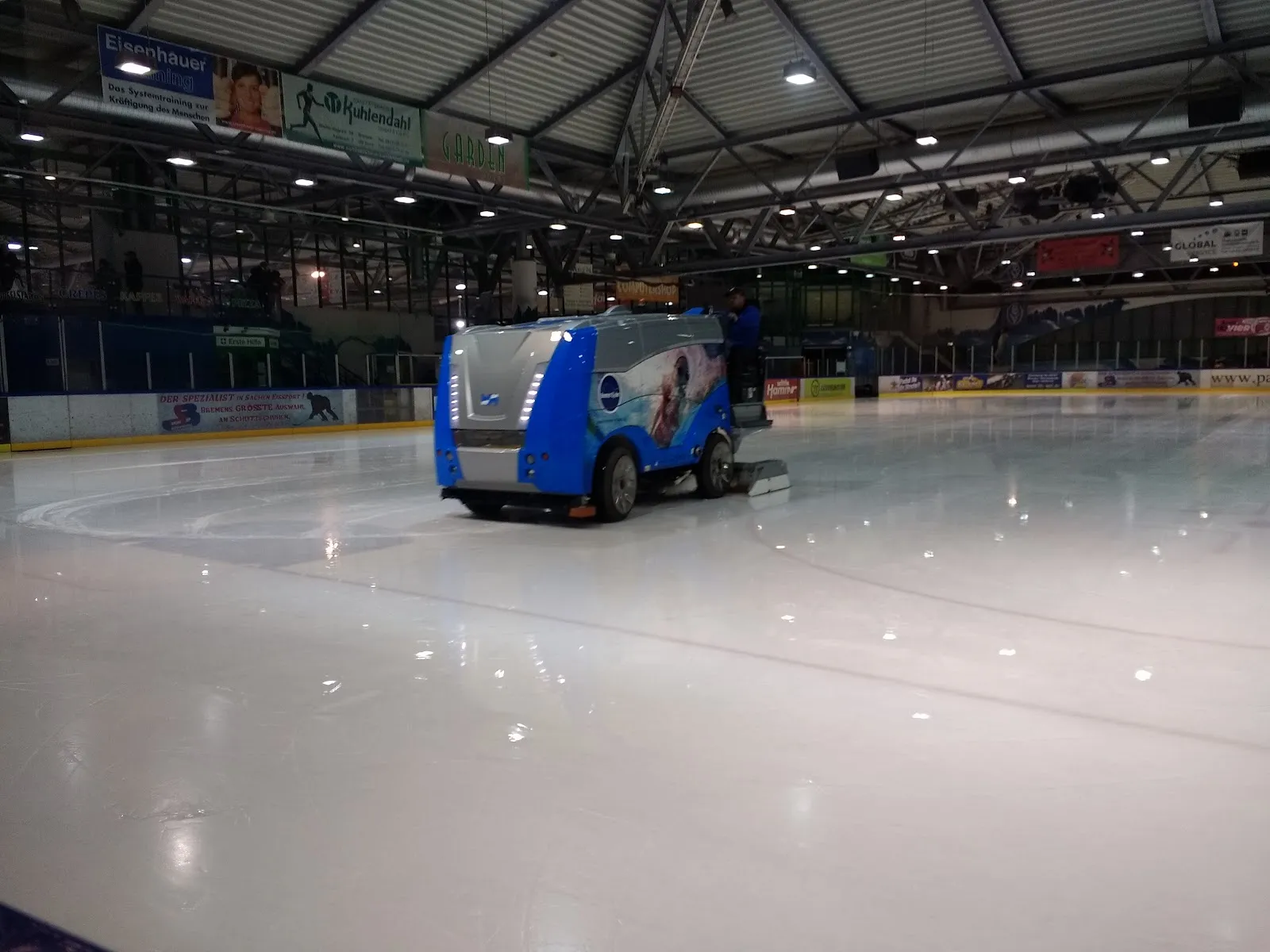 Die Eisfläche der Halle Paradice, frisch von der blauen Eisbereitungsmaschine abgezogen, mit Banden und Halle dahinter; keine Personen. – Eissporthalle Paradice