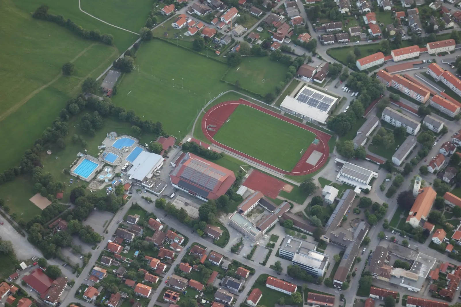 An aerial view of the ice hall's roof beside the running track and the open-air pool, with the town around it. — Eissporthalle Peißenberg