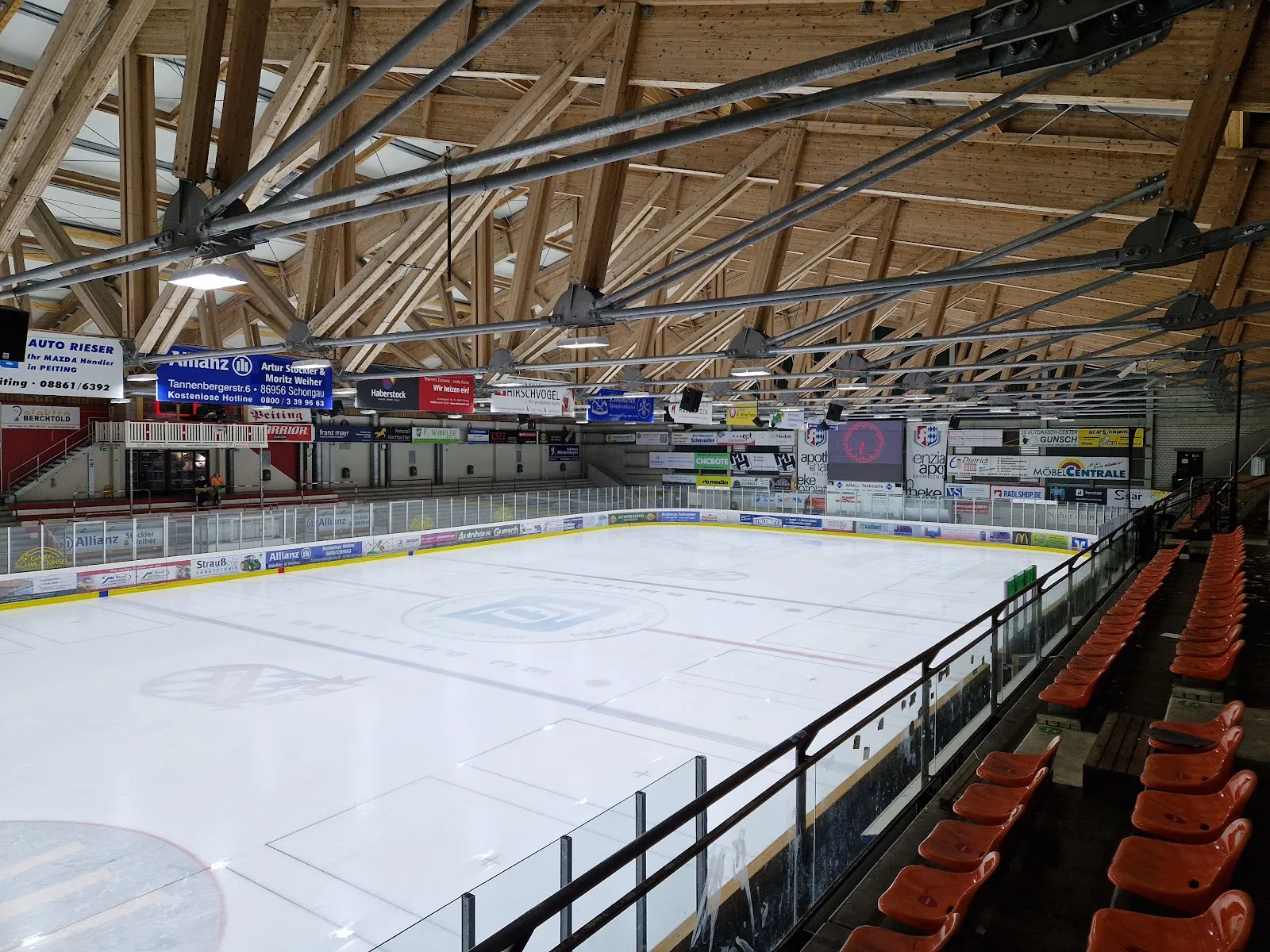 The hall's ice surface seen from the stands, with the orange seats at the right, the glazed boards and the timber roof beams above. — Eissporthalle Peißenberg