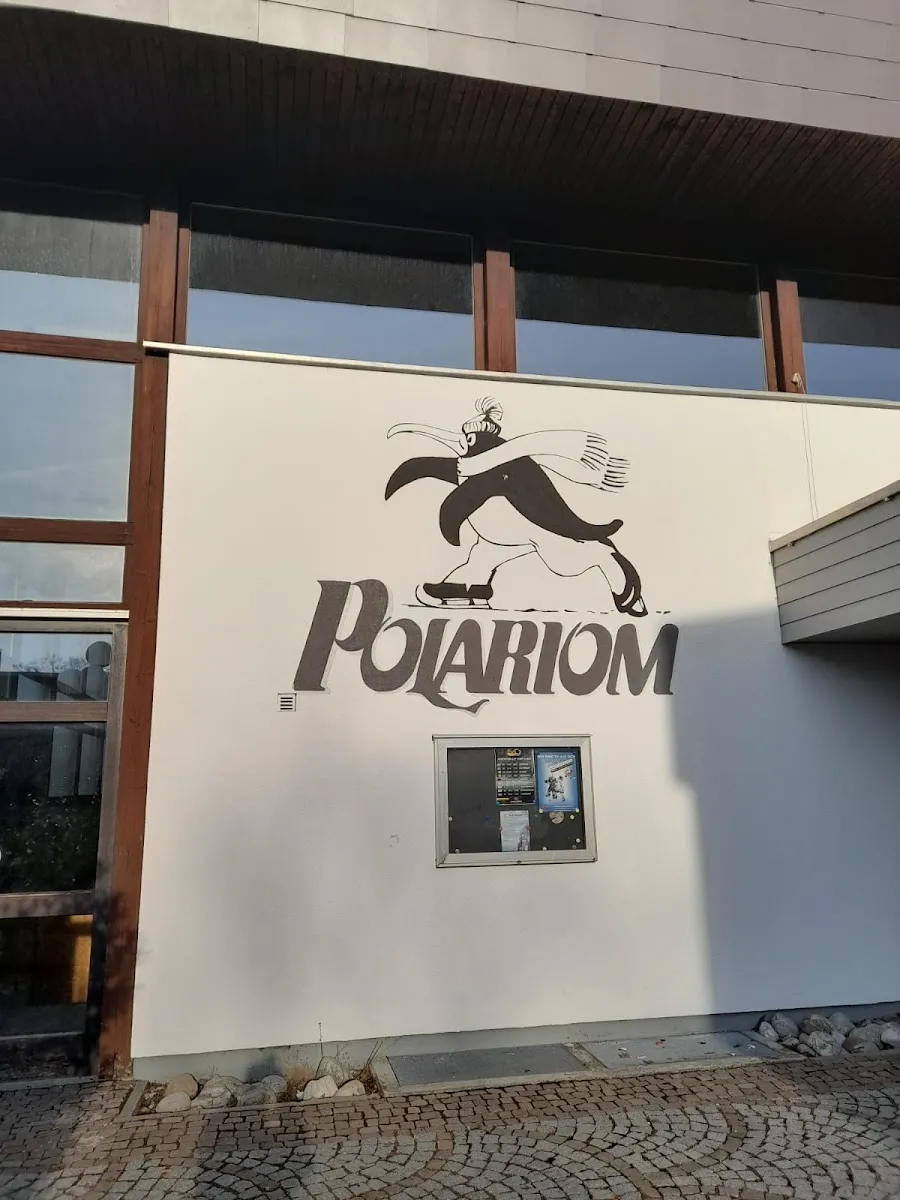 The white wall at the entrance with the penguin-on-skates Polariom logo, glazed bays and a cobbled apron below. — Eissporthalle Polariom Germering