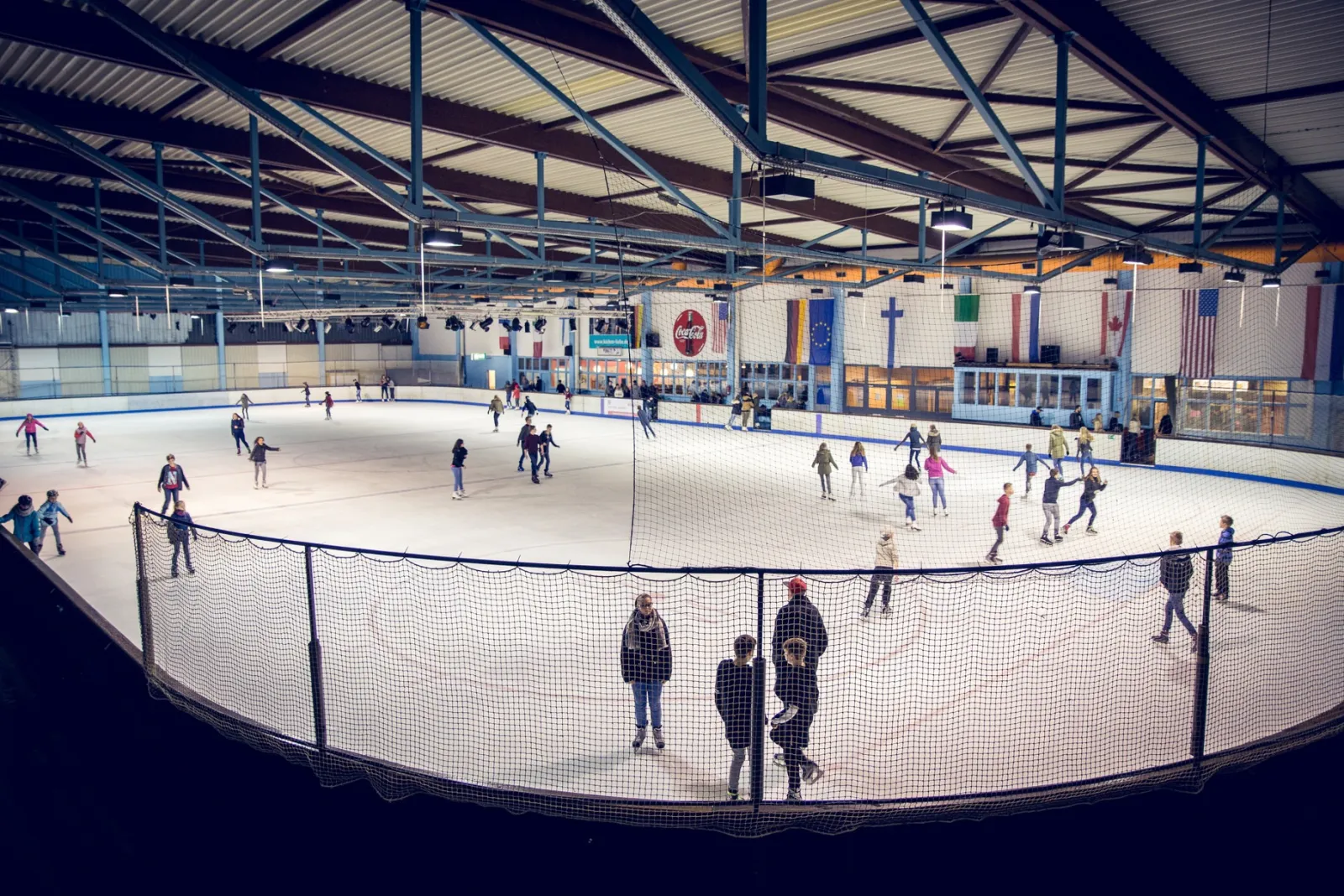 The Rheine ice hall during a public session, skaters spread over the ice and the roof trusses and glazed side wall overhead. — Eissporthalle Rheine
