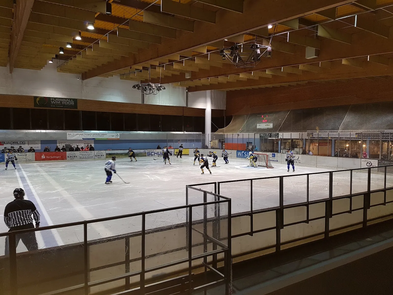 Ein laufendes Spiel auf der Eisfläche in Solingen, Spieler über dem Eis unter dem Holzdach, im Vordergrund das Bandenglas. – Eissporthalle Solingen