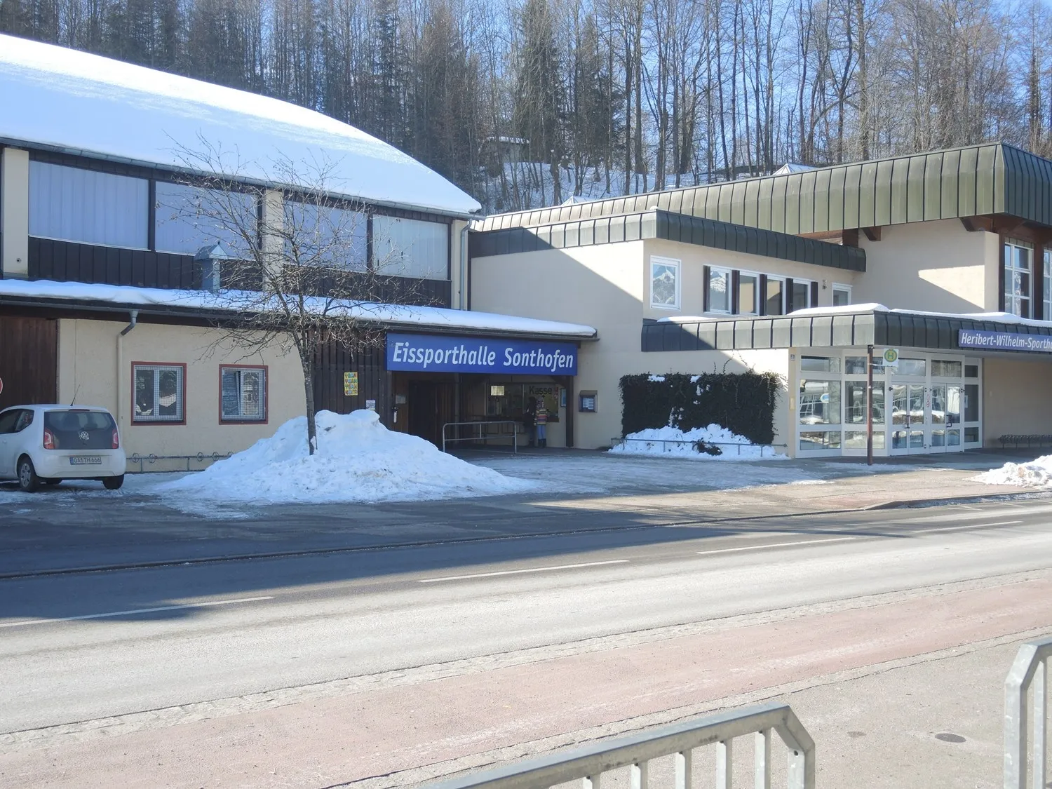 The entrance front of the Eissporthalle Sonthofen on a sunny winter day, with the blue Eissporthalle Sonthofen sign above the doors, the arched hall roof and snow along the road. — Eissporthalle Sonthofen