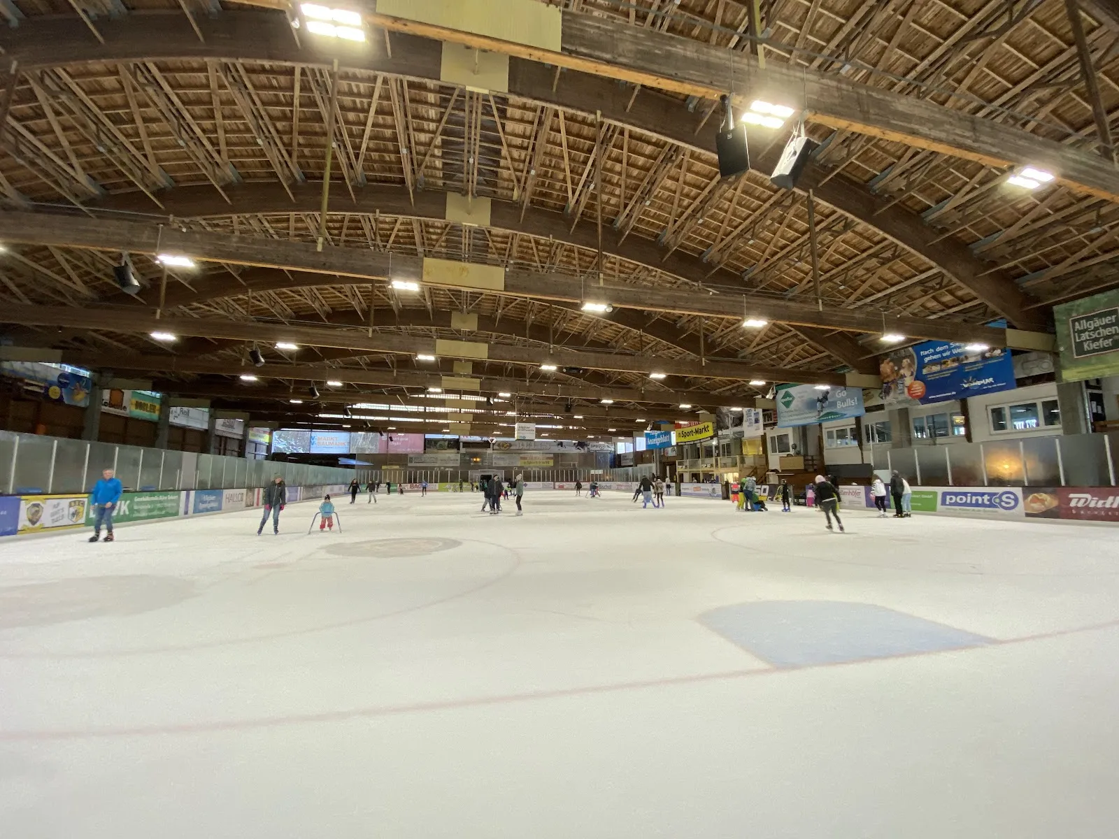 A wide view along the ice sheet under the hall's timber roof structure, with a few skaters in the distance and sponsor boards along the sides. — Eissporthalle Sonthofen