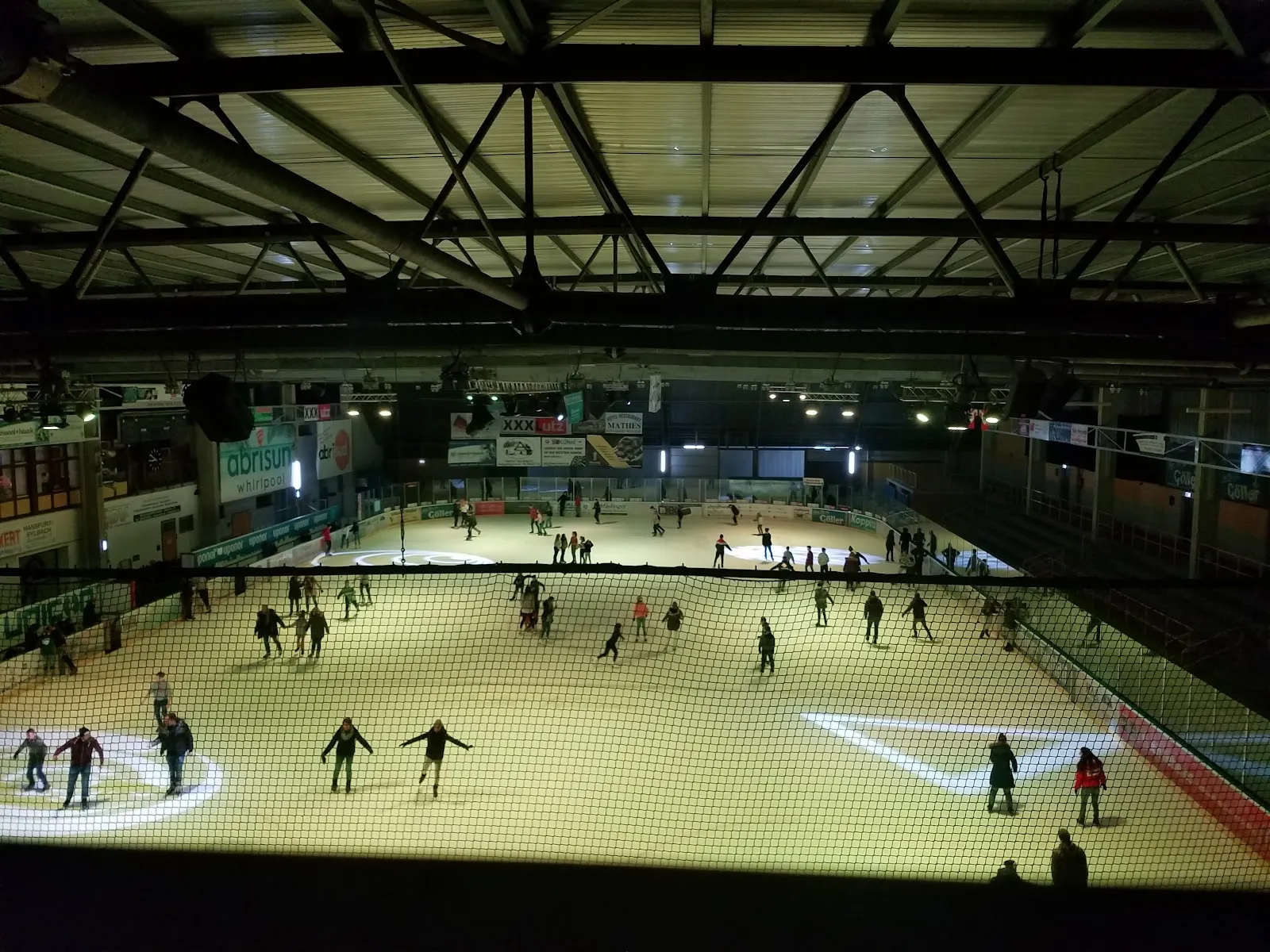 Ein abendlicher Publikumslauf, von den Rängen über ein Netz hinweg gesehen, mit der beleuchteten Eisfläche unter Stahlbindern im Dach und Werbebanden an den Wänden. – Eissportstadion Haßfurt